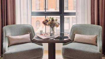 Duas poltronas viradas uma para a outra, com uma mesa no meio, um vaso de flores e uma janela com vista para um edifício de tijolos.