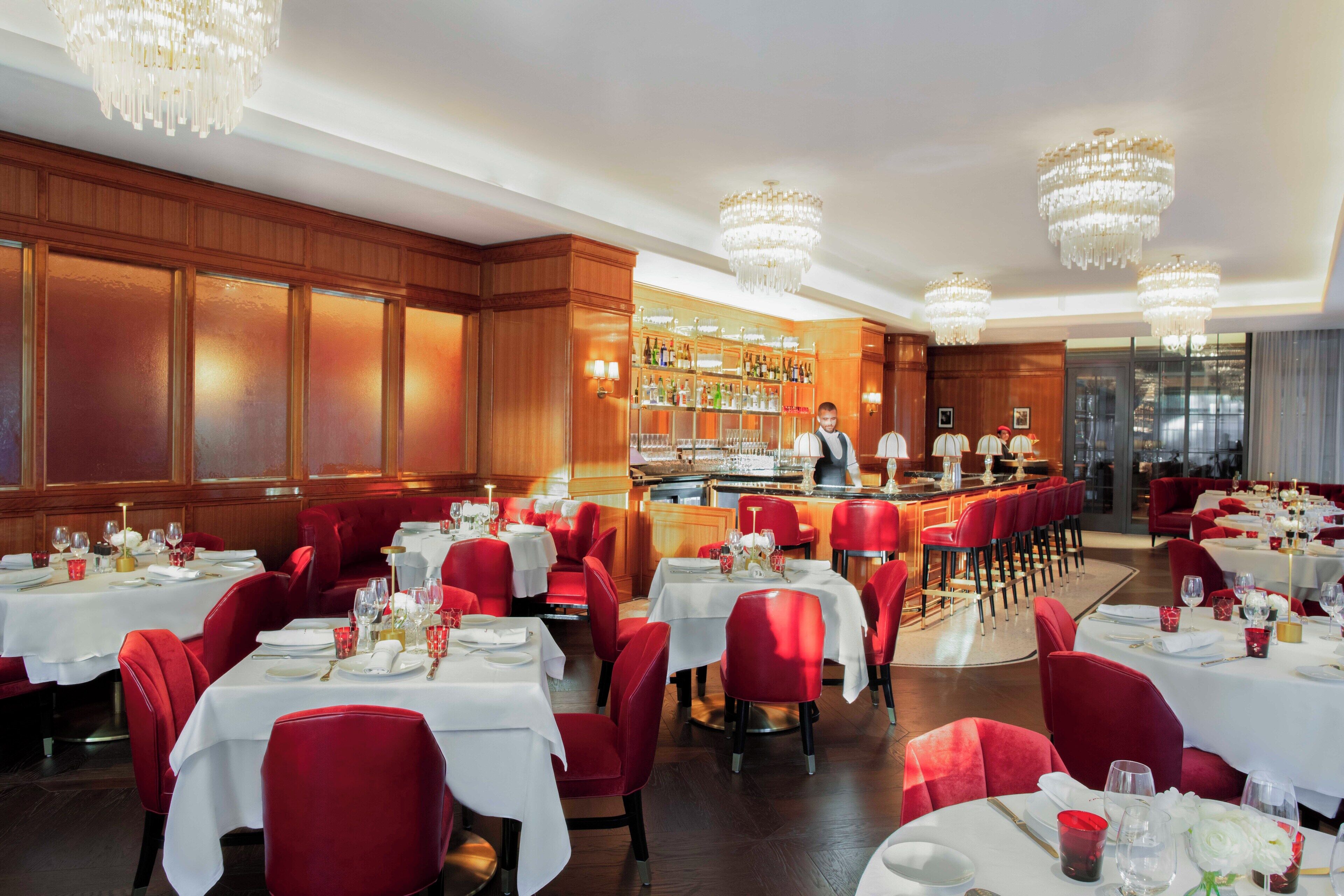A restaurant with red upholstered chairs, white tablecloths, and a bar area.