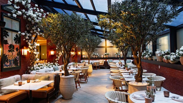 Un área de comedor interior y exterior con mesas y sillas, plantas en macetas y una pared de ladrillo.