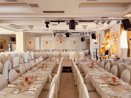 Un salón de banquetes con largas mesas dispuestas para un evento formal, con candelabros, luces de escenario y un arco de globos decorativo.