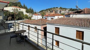 Un balcon avec une table et des chaises, donnant sur une ville aux bâtiments aux toits de tuiles et sur une colline en arrière-plan.