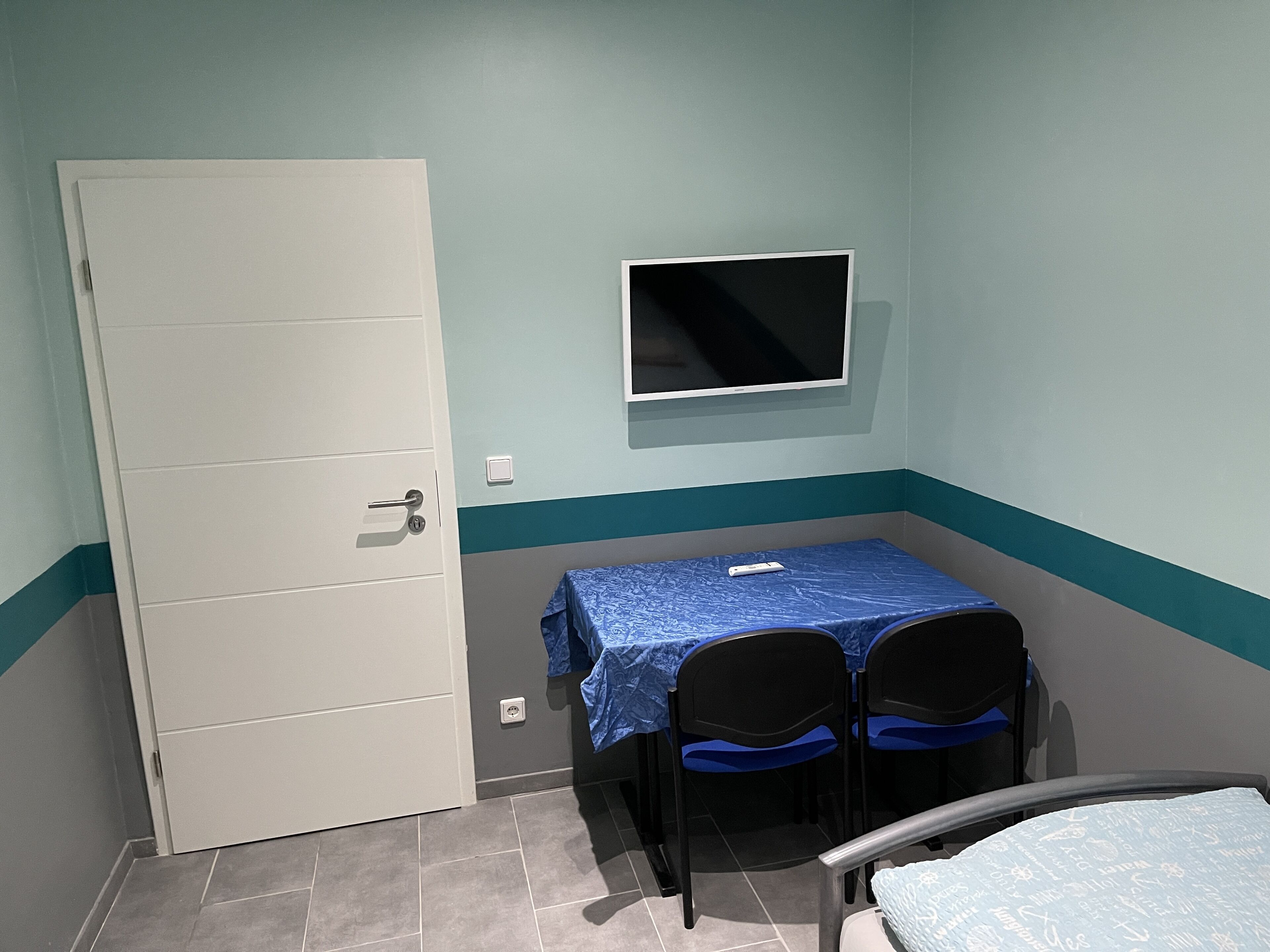 Una habitación pequeña con una cama individual, una mesa con dos sillas, un televisor en la pared y una puerta.