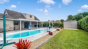 Ein Wohnpool mit überdachter Terrasse, einer kleinen Gartenhütte und einem gepflegten Rasen.
