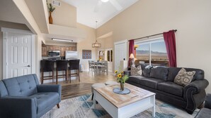A spacious living room with a gray sofa, a wooden coffee table, and a view of the mountains through a large window. - The Oasis - Golf - Pool - Hot Tub - Condo with a View. (Mesquite)