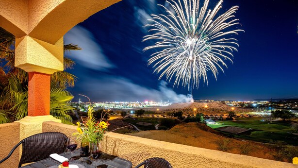 A table set for two with a dessert, a vase with flowers, and a view of fireworks over a cityscape. - The Oasis - Golf - Pool - Hot Tub - Condo with a View. (Mesquite)
