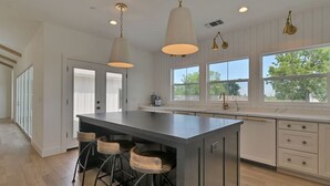 Una cocina moderna con una isla grande, taburetes de bar, iluminación colgante y ventanas amplias.