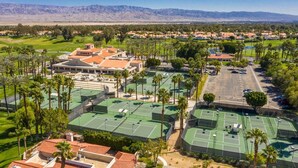 Un gran complejo deportivo con varias canchas de tenis, rodeado de edificios residenciales y palmeras.