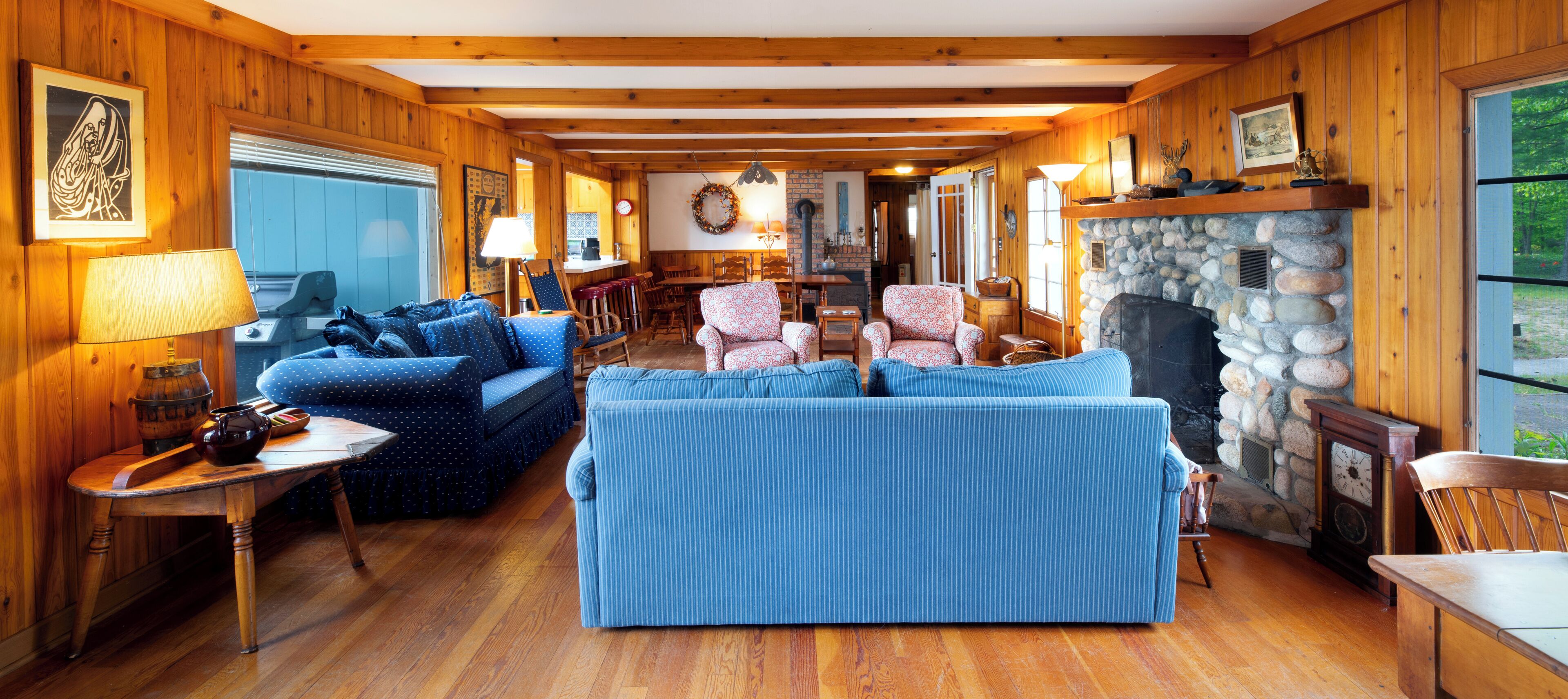L’intérieur chaleureux d’un chalet avec des murs en bois, une cheminée en pierre, un coin repas et un salon doté de canapés à rayures bleues.