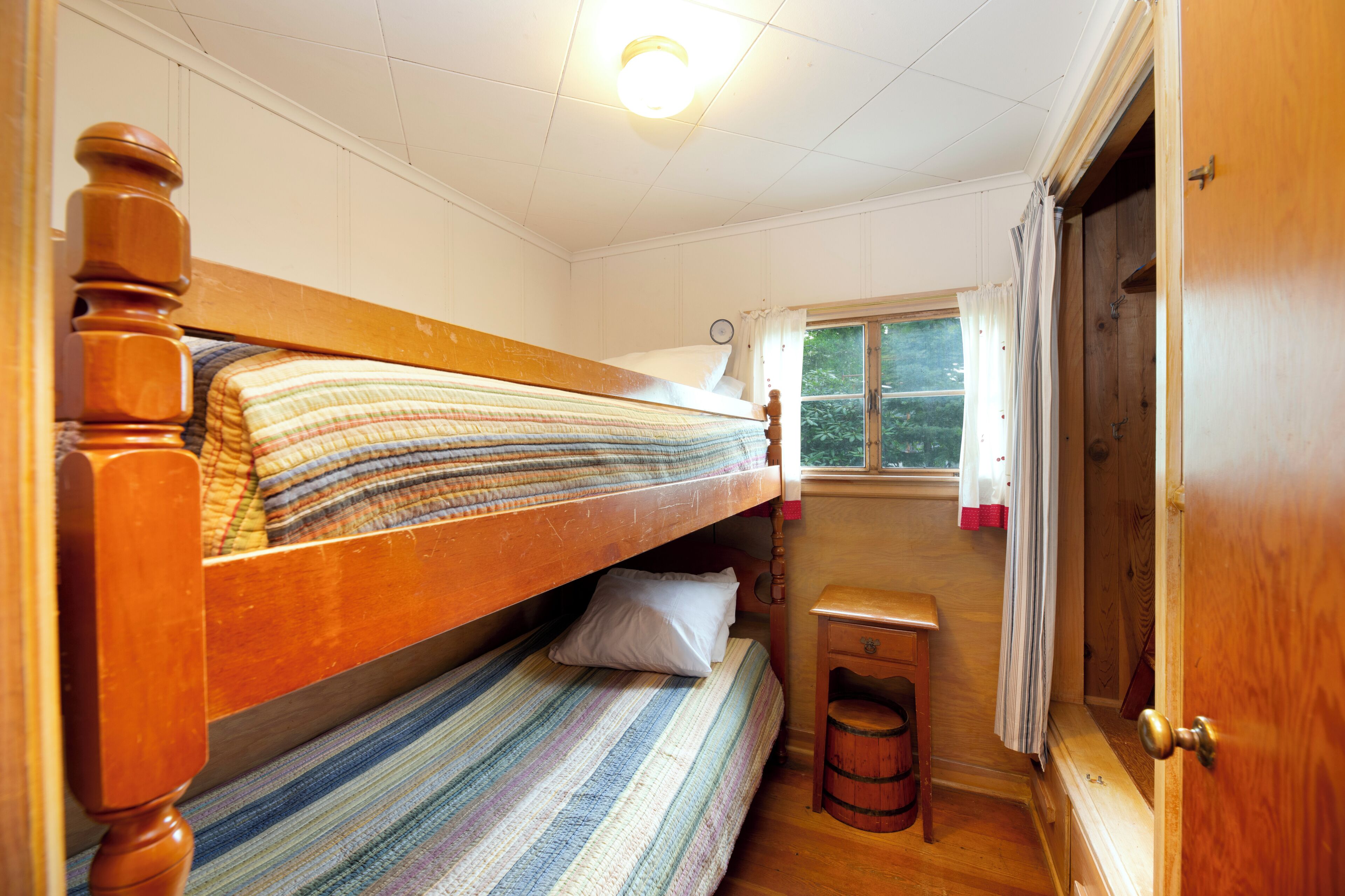 A room with two wooden bunk beds, a small wooden stool, a window with curtains, and a door.