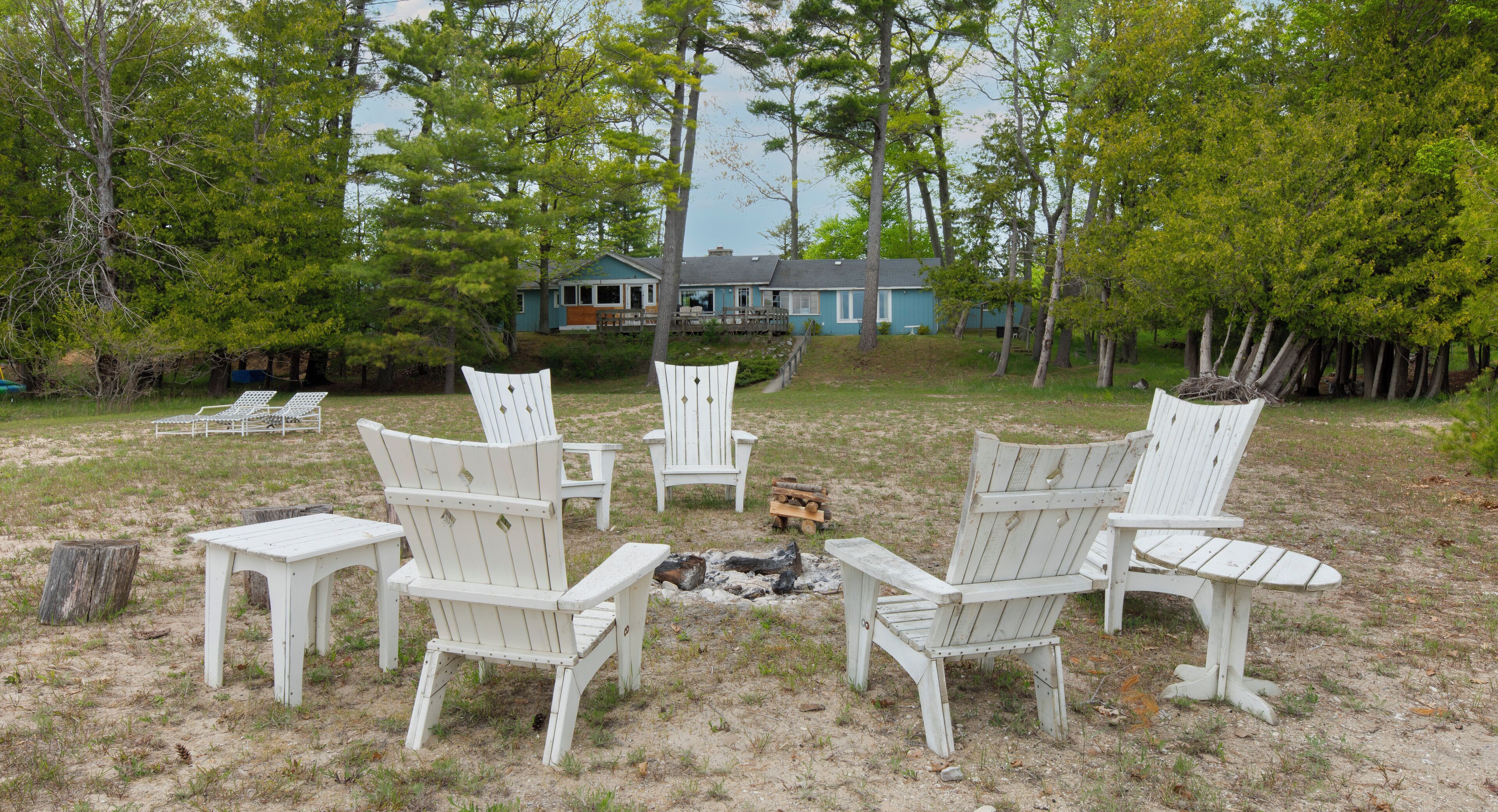 A group of white Adirondack chairs arranged in a circle around a small fire pit in a grassy area with trees and a house in the background.