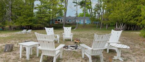 A group of white Adirondack chairs arranged in a circle around a small fire pit in a grassy area with trees and a house in the background.