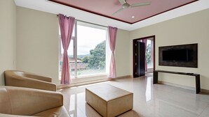 A spacious living room with a large window, a flat-screen TV mounted on the wall, and a ceiling fan. - Itsy Hotels Kattari Komforts, Chikmagalur (Chikkamagaluru)