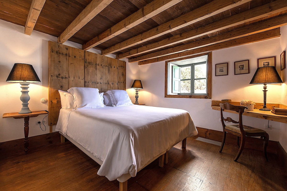 Ein Schlafzimmer mit Holzboden, einem Bett mit weißen Leinentextilien, zwei Nachttischen mit Lampen, einem Fenster und gerahmten Bildern an der Wand.