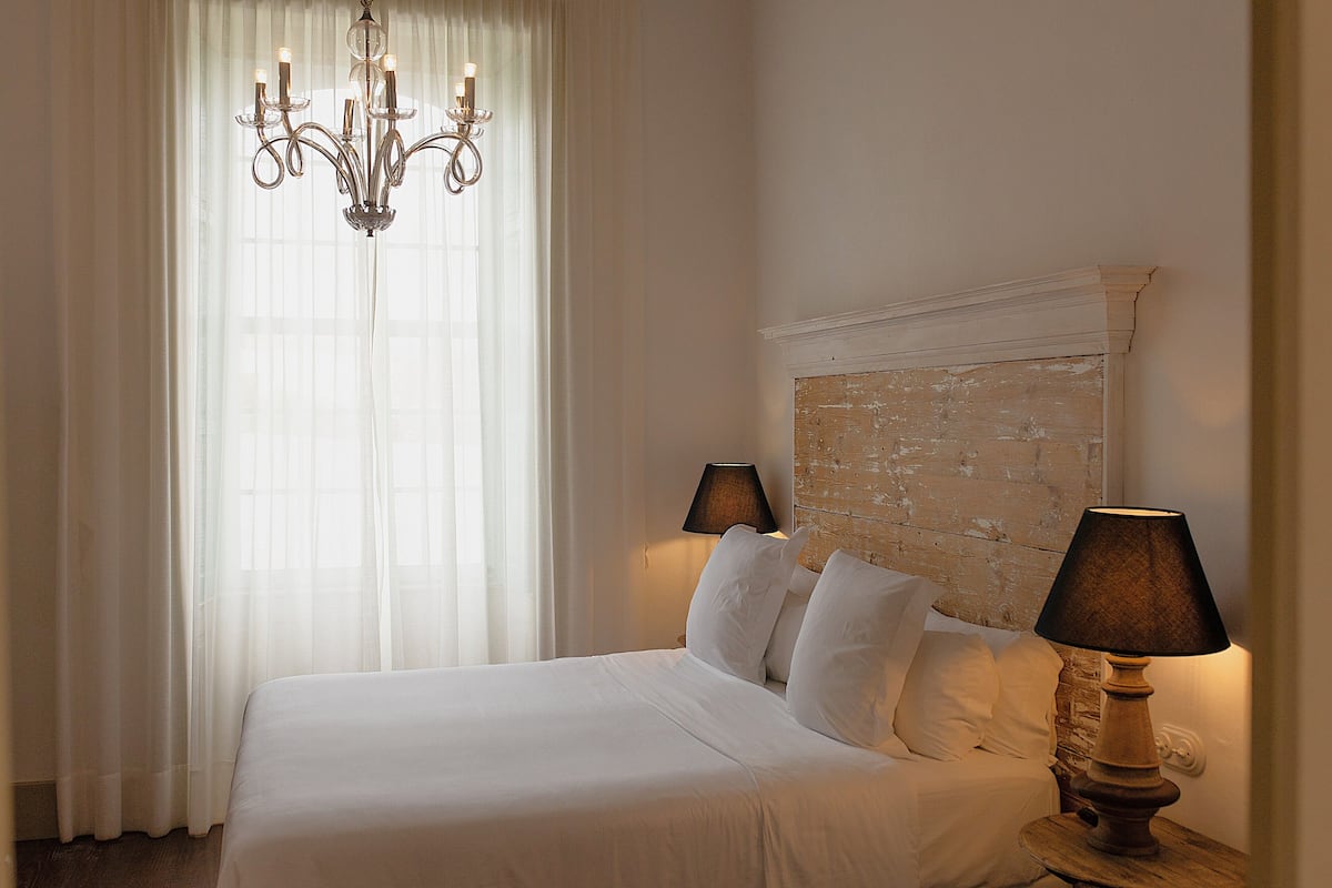 Ein Hotelzimmer mit einem großen Bett, zwei Nachttischlampen, einer Deckenleuchte und einem Fenster mit durchsichtigen Vorhängen.