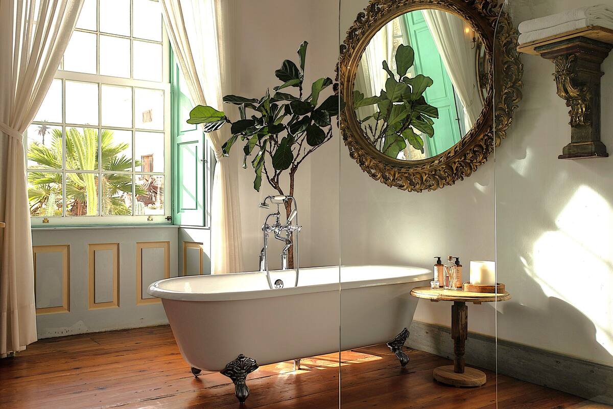 Ein Badezimmer mit freistehender Badewanne, einem Spiegel und einem fensterförmigen Ausblick mit Vorhängen.