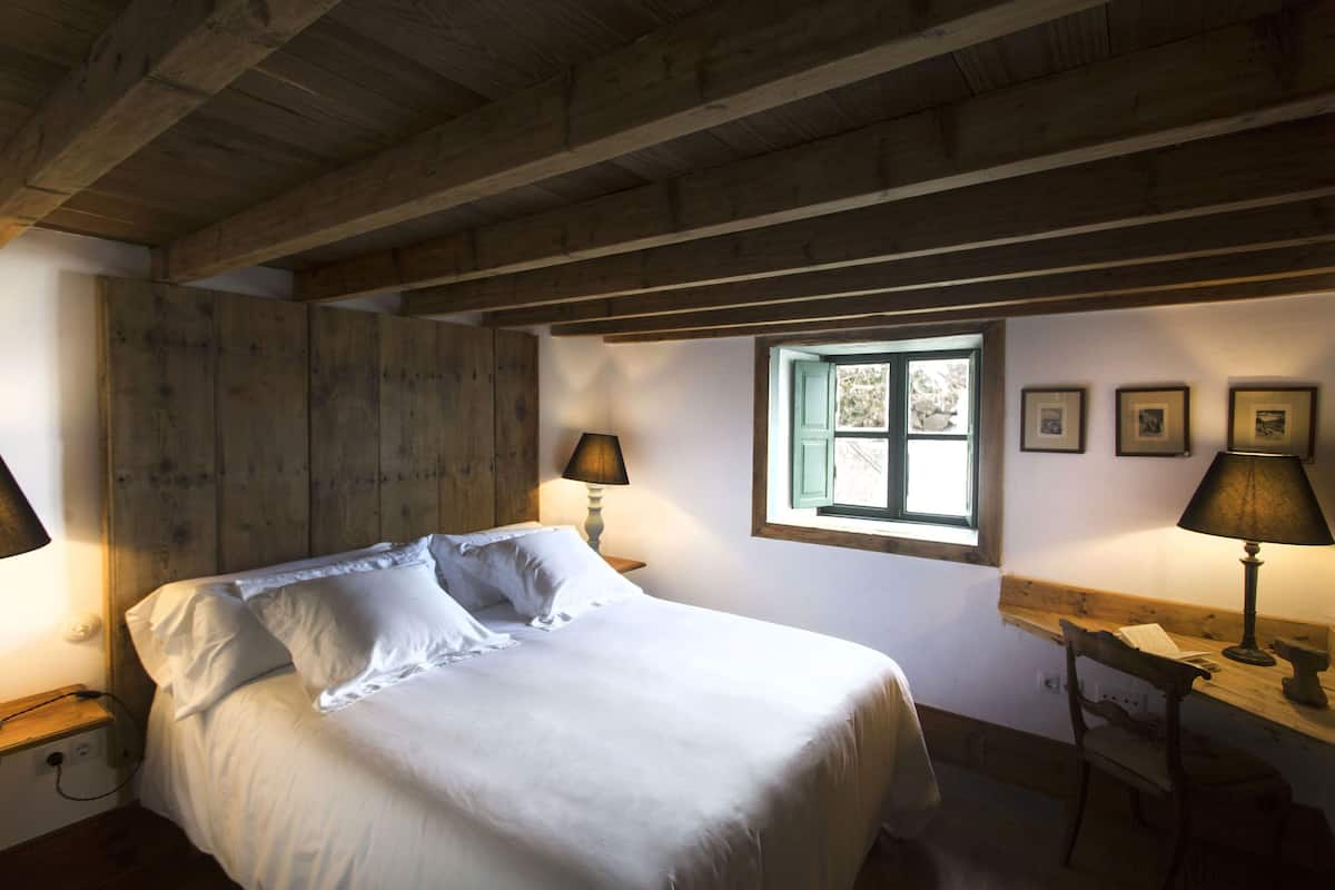 Ein Schlafzimmer mit Holzboden, einem Bett mit weißen Bettwäsche, zwei Nachttischlampen, einem Fenster mit Aussicht und einem Schreibtisch mit Lampe.