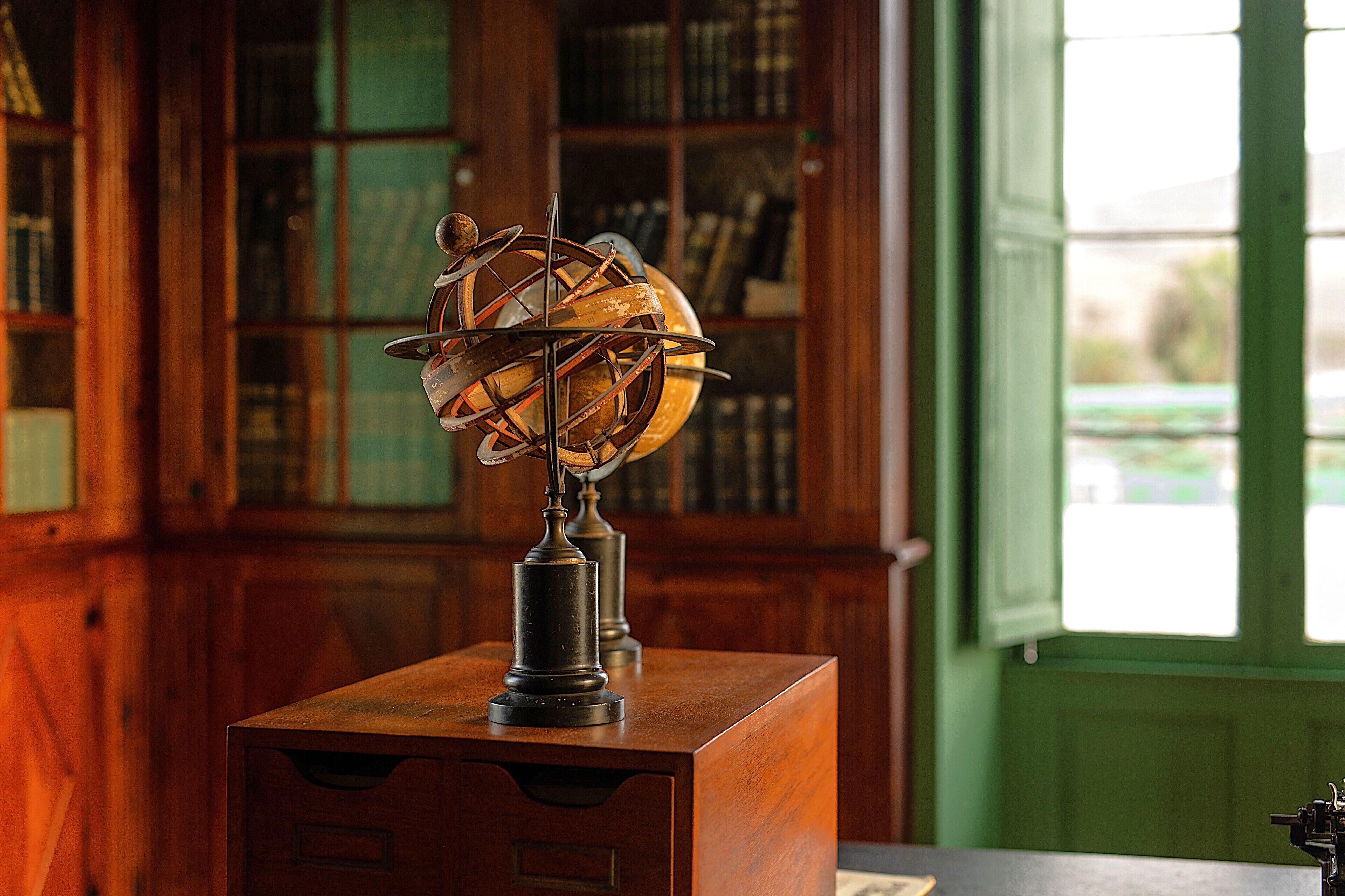A globe on a wooden desk in a room with wooden bookshelves and a window.