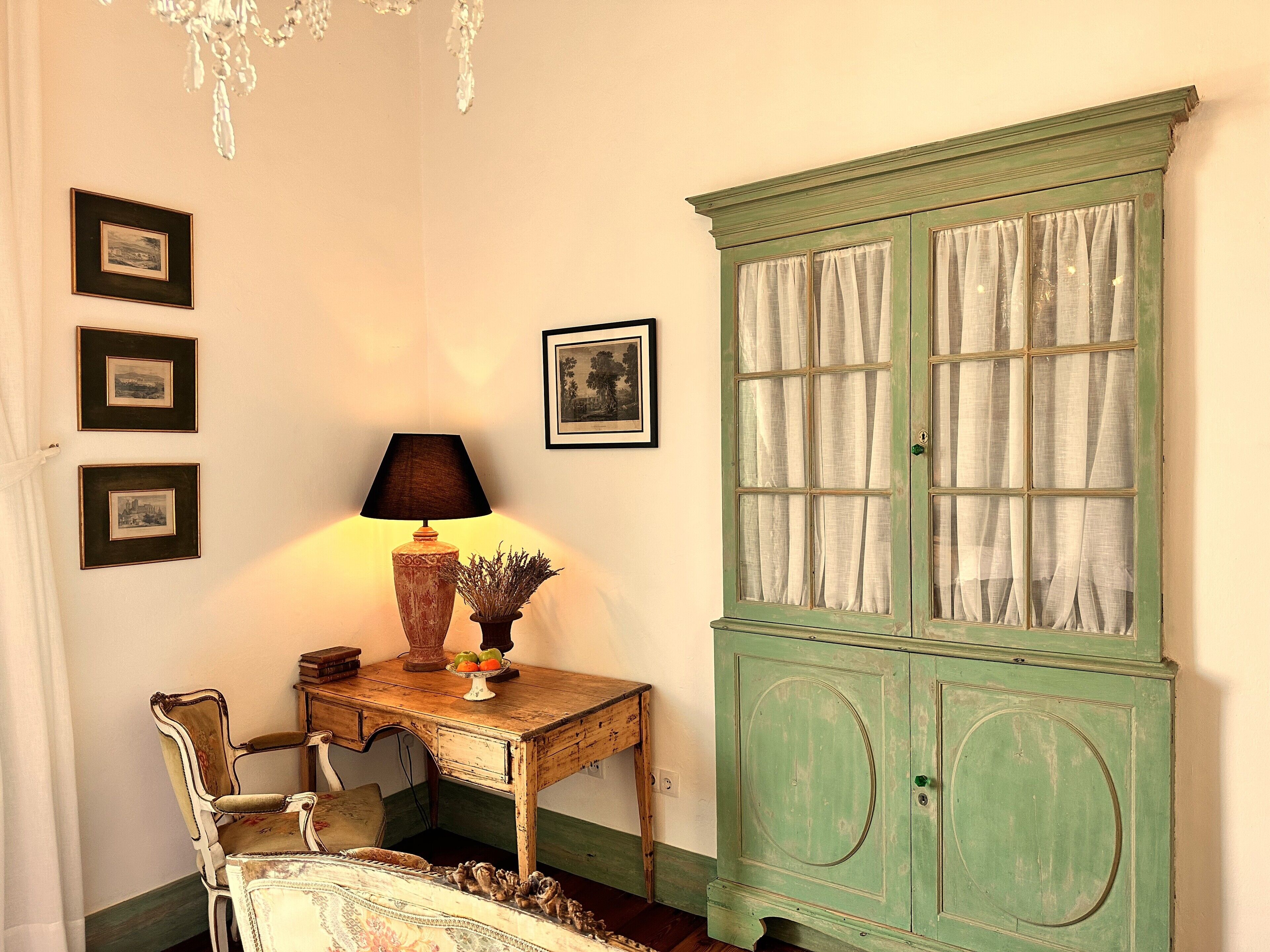 Een kamer met een groen kastje, een houten bureau met een lamp, een stoel en beelden in lijsten aan de muur.