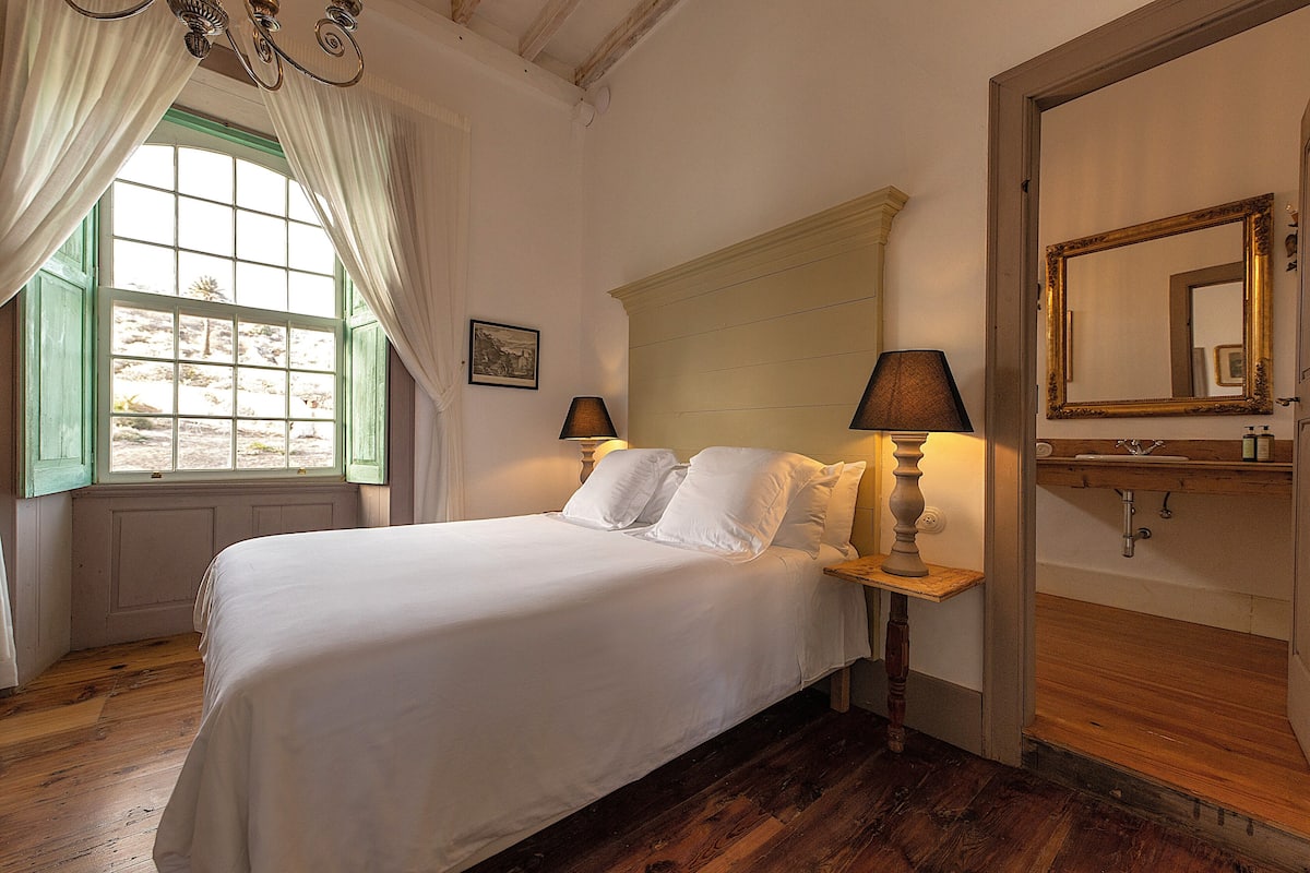 Ein Schlafzimmer mit einem großen Bett, einem Nachttisch mit Lampe, Holzboden, einem Fenster mit Vorhängen und einem Spiegel an der Wand.