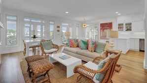 A spacious living room with a sofa, coffee table, and wicker chairs, a dining area with a table and chairs, and a kitchen with white cabinets.