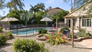 Un patio trasero con piscina, rodeado por una cerca, con palmeras, un flotador de flamenco y una zona de patio con sombrillas.