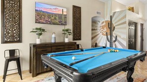 Una mesa de billar con bolas, un televisor, paneles decorativos y un mural con motivos de cactus en una habitación que tiene una puerta y una planta.