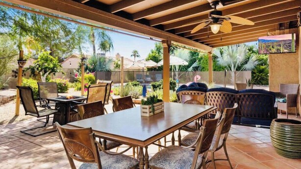 Un patio al aire libre con mesa y sillas de madera, un ventilador de techo y vistas a un jardín con palmeras y una piscina.
