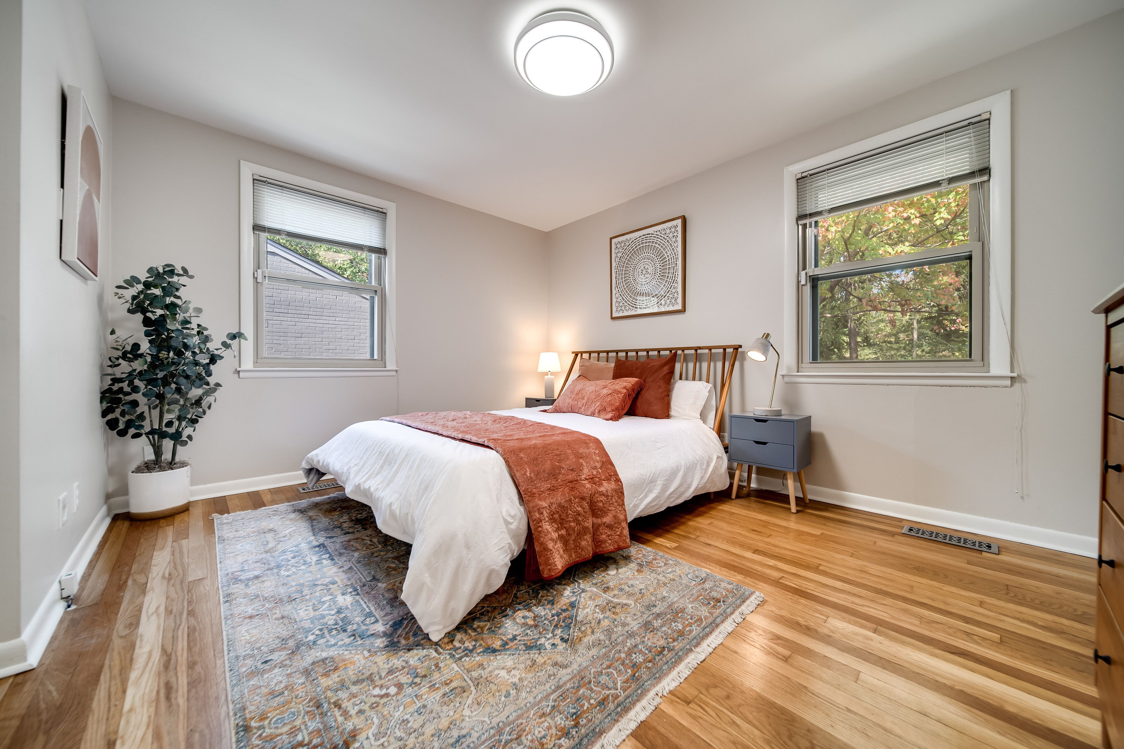 A bedroom with a bed, a nightstand, a lamp, a potted plant, and a window with blinds.