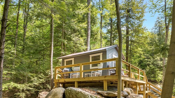 A cabin with a wooden deck and a covered outdoor seating area, surrounded by a forest. - Cozy Cottage overlooking stream on 60 acres (Middle Grove)