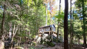 A wooden cabin with a deck, surrounded by dense forest and rocky terrain. - Cozy Cottage overlooking stream on 60 acres (Middle Grove)