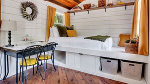 A cozy room with a bed, a desk, and chairs. There is a window with curtains, a wall-mounted shelf with books, and a decorative wreath. - Cozy Cottage overlooking stream on 60 acres (Middle Grove)