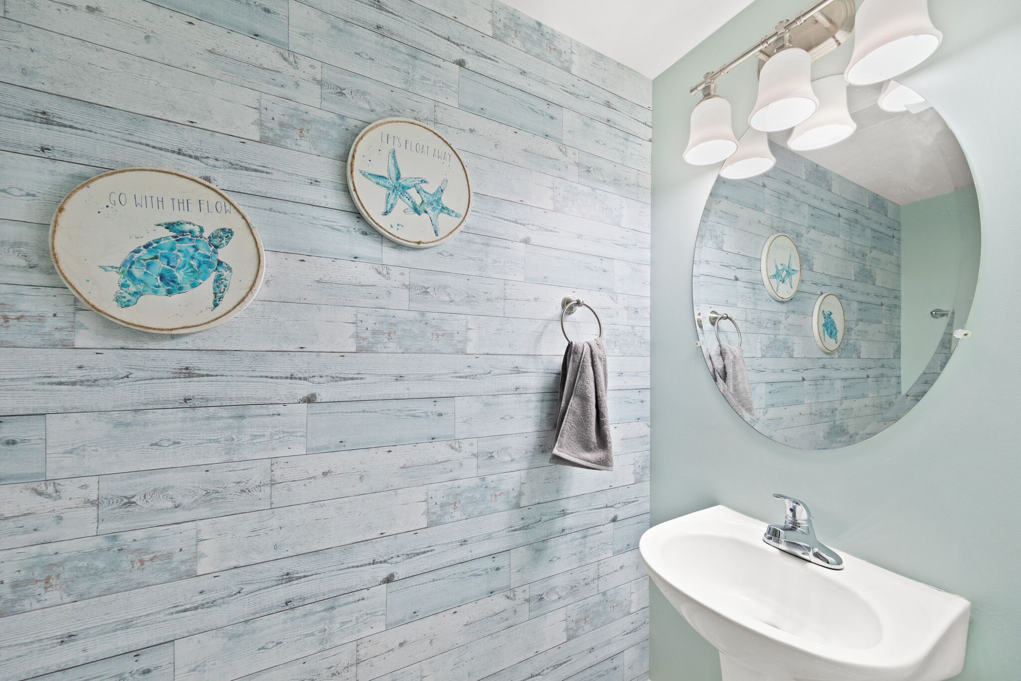 A bathroom with a wooden wall, a round mirror, a sink, and two decorative plates with sea turtle and starfish designs.