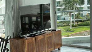A modern living room with a large flat-screen TV on a wooden cabinet, a dining area with a table and chairs, and a view of a well-maintained garden and building outside. - CONDO FLAMINGO  BY TRVL2HM (Nuevo Nayarit)