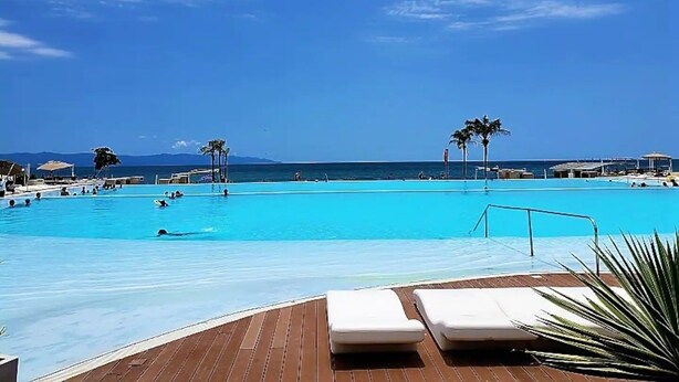 A large outdoor swimming pool with clear blue water, surrounded by lounge chairs and palm trees, with a view of the ocean and mountains in the background. - CONDO FLAMINGO  BY TRVL2HM (Nuevo Nayarit)