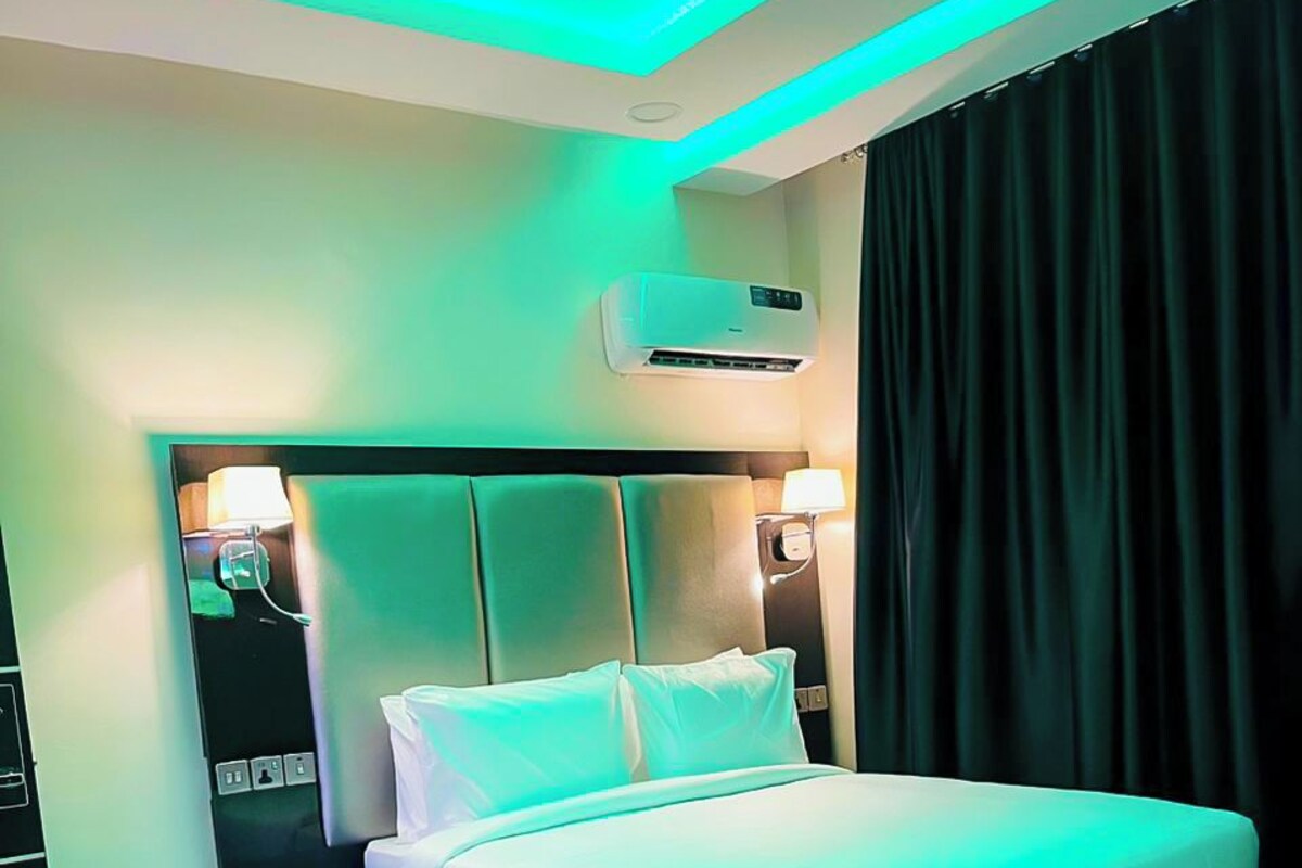 A hotel room with a large bed, bedside tables, a desk, and an air conditioning unit.