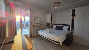 A bedroom with a bed, a desk, a chair, a wardrobe, and a balcony with a view.