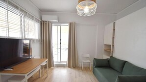 A modern living room with a flat-screen TV, wooden flooring, a green sofa, a white chair, and a pendant light.