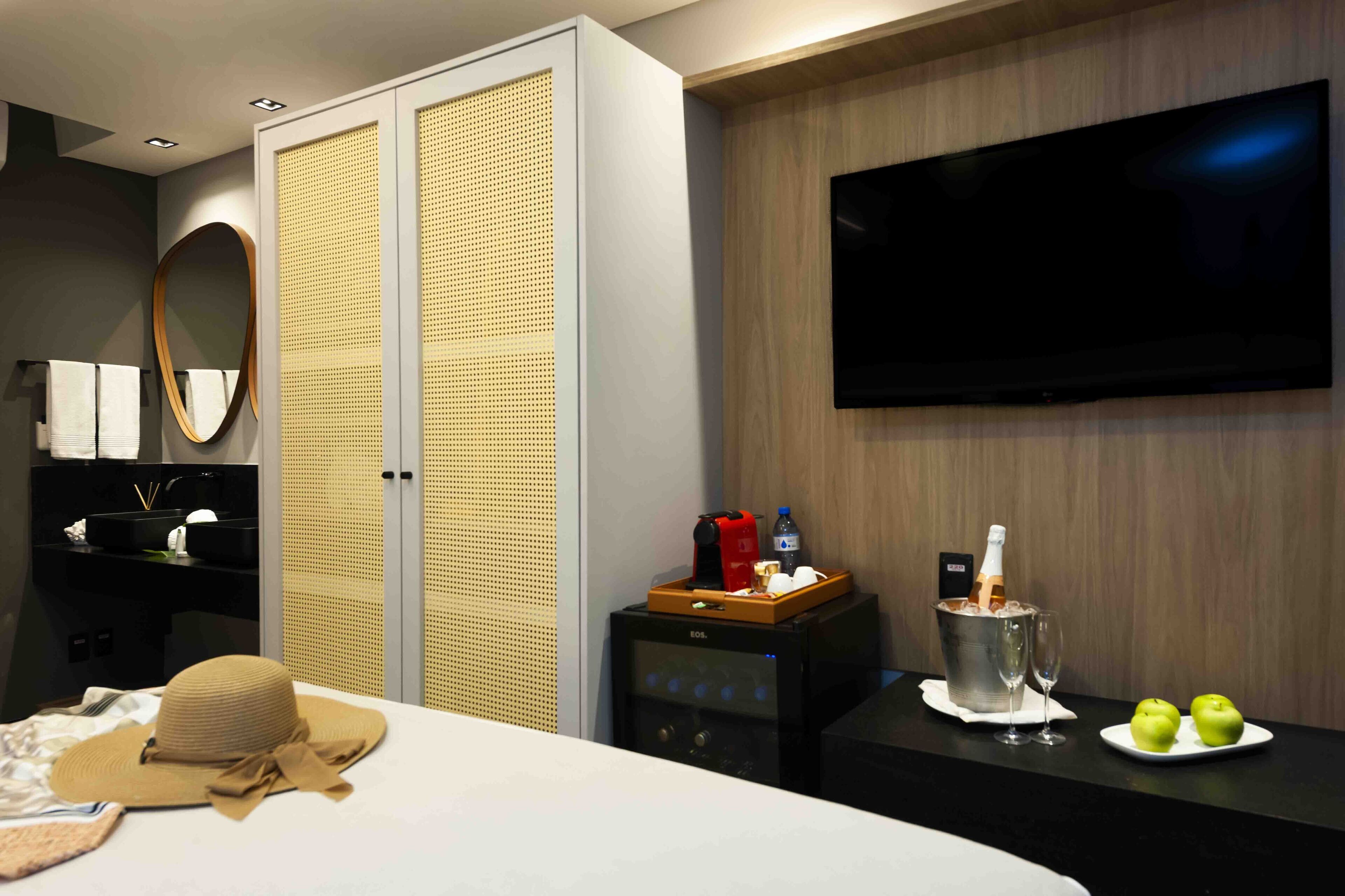 A hotel room with a bed, a TV mounted on the wall, a minibar, and a small table with a bottle of champagne and two apples.