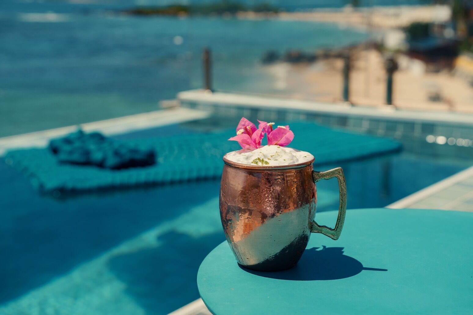 Una taza de cobre con adorno floral sobre una mesa turquesa junto a una piscina.