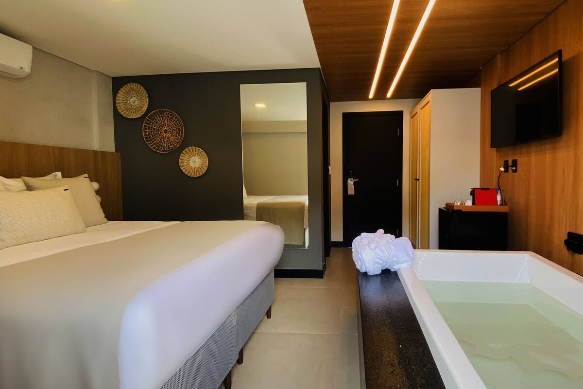 Ein Hotelzimmer mit Bett, einem Badezimmer inklusive Badewanne, einem Spiegel und einem an der Wand montierten Fernseher.