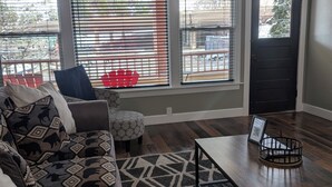 A living room with a sofa, ottoman, coffee table, and large windows with blinds. - Cozy, remodeled townhome retreat in downtown Historic Wallace! (Wallace)