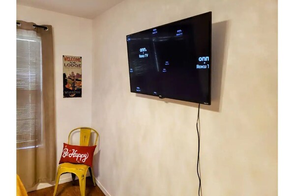 A room with a mounted TV displaying 'onn Roku TV' and 'onn Roku 1'. A yellow chair with a 'Be Happy' pillow is visible.