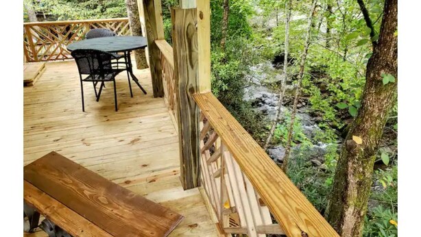 Une terrasse en bois, agrémentée d’une table et de chaises, entourée d’une balustrade en bois, et offrant une vue sur une zone boisée traversée par un ruisseau.