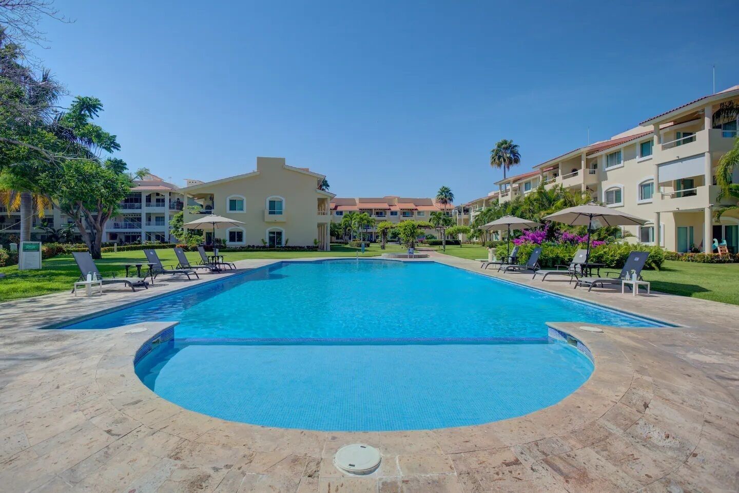 Piscina com espreguiçadeiras, cercada por área ajardinada com palmeiras e um edifício com várias varandas.