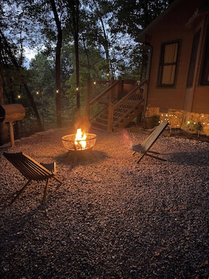 A fire pit with a wooden bench and a lounge chair in a gravel area, surrounded by trees and a building with a staircase. - Luxury Cabin w/ views•Toccoa•<br> HotTub•Firept•Pets (Blue Ridge)