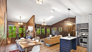 Un vaste espace de vie avec un intérieur en bois, une table à manger entourée de chaises blanches, un canapé et une cuisine équipée d’appareils électroménagers modernes.