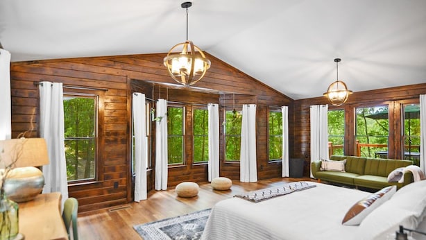 Une chambre spacieuse aux murs en bois, dotée d’un grand lit, de deux luminaires suspendus et offrant une vue sur la verdure environnante.