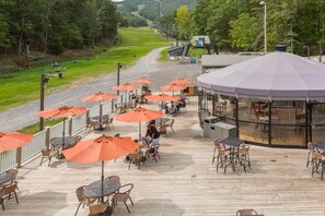 Eine Terrassenfläche im Freien mit Tischen und orangefarbenen Sonnenschirmen, mit Blick auf eine Straße und Grünflächen.
