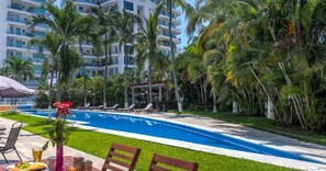 Piscina com espreguiçadeiras, palmeiras e um edifício ao fundo.