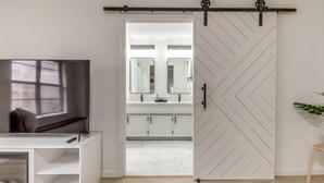 A modern bathroom with a sliding door, a vanity with a mirror, and a television on a stand. - Chic OKC Retreat Near Lake Hefner -Family Friendly (Oklahoma City)